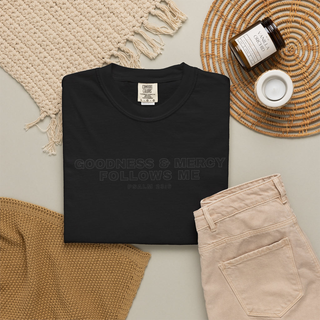 Black t-shirt on a light surface with Goodness and Mercy Follow Me Psalm 23:6 with beige pants and decorative items.