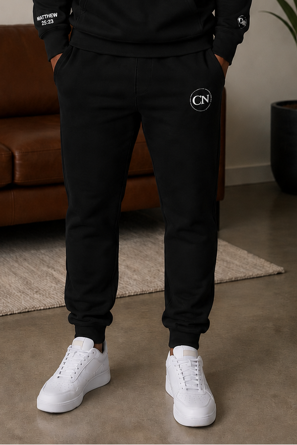 Man wearing the Courageous Nation Faithful Everyday Jogger Set in black, standing confidently in a modern living room with faith-inspired wall art in the background