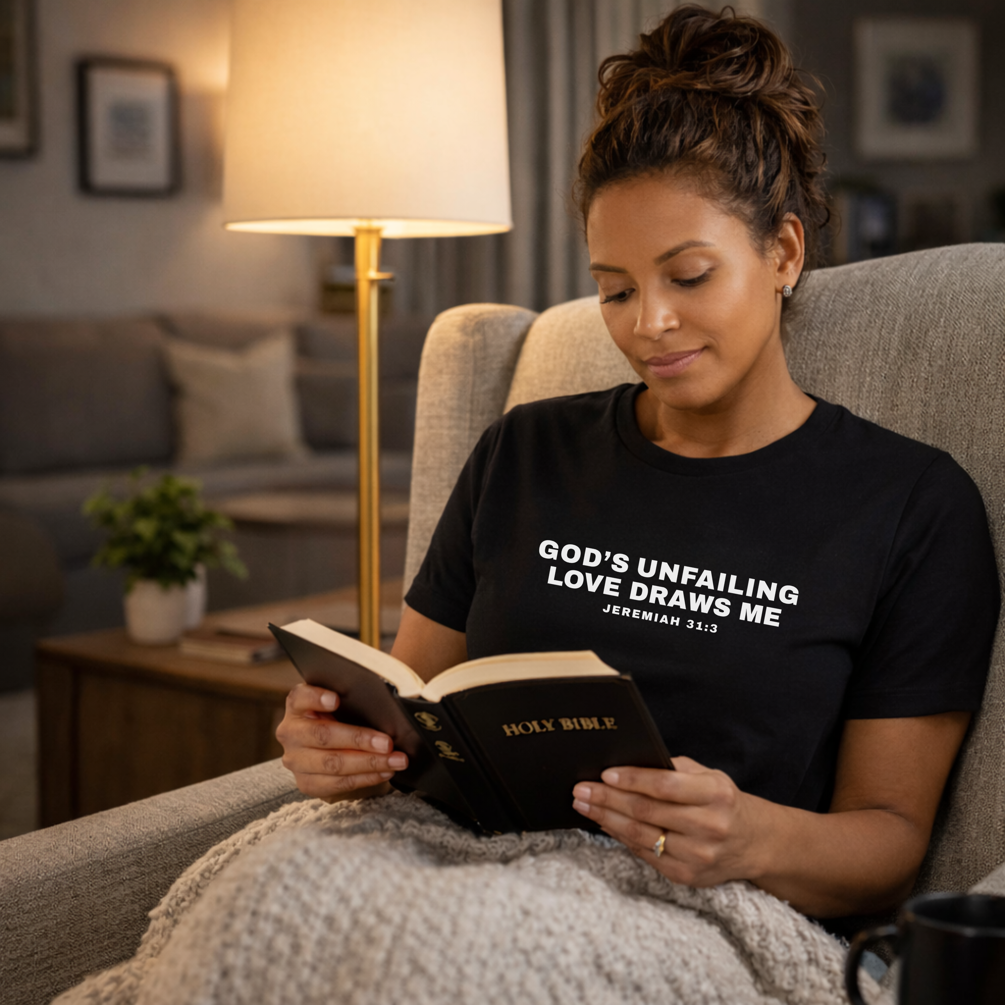 Woman reading the Holy Bible in a cozy living room wearing a black t-shirt with that says; God's Unfailing Love Draws Me Jeremiah 31:3.