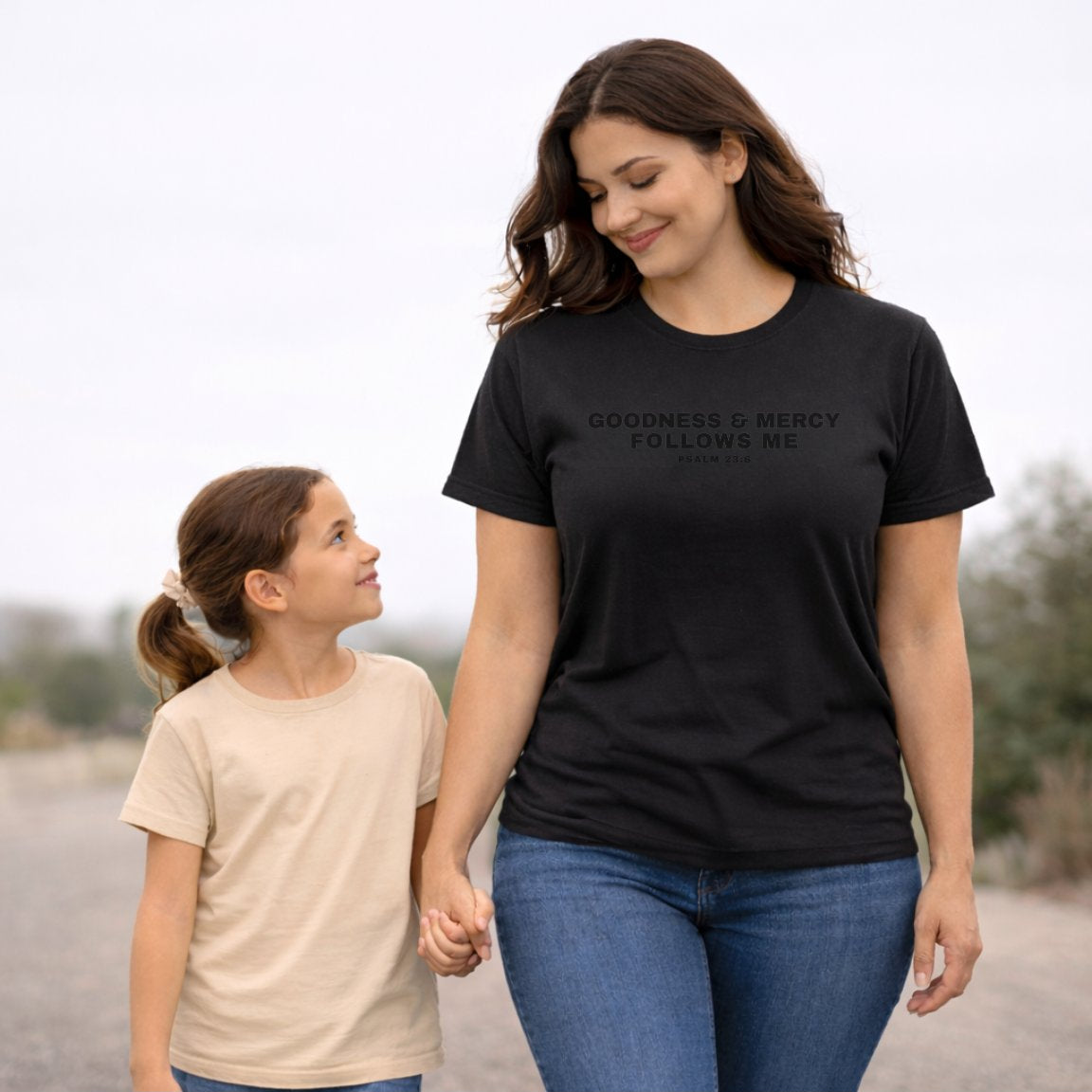 Woman wearing a Goodness And Mercy Follows Me Psalm 23:6 t-shirts and child holding hands outdoors.