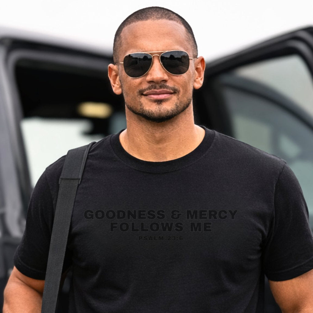 Man wearing a black t-shirt with Goodness and Mercy Follows Me Psalm 23:6, standing in front of a vehicle.